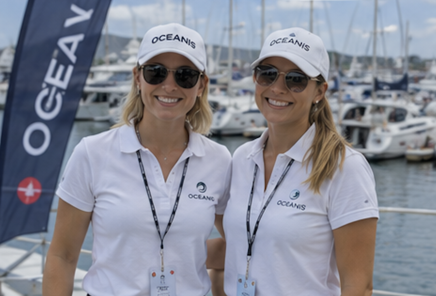 Two female hospitality hosts in white branded caps and polos smiling in front of boats at a marina