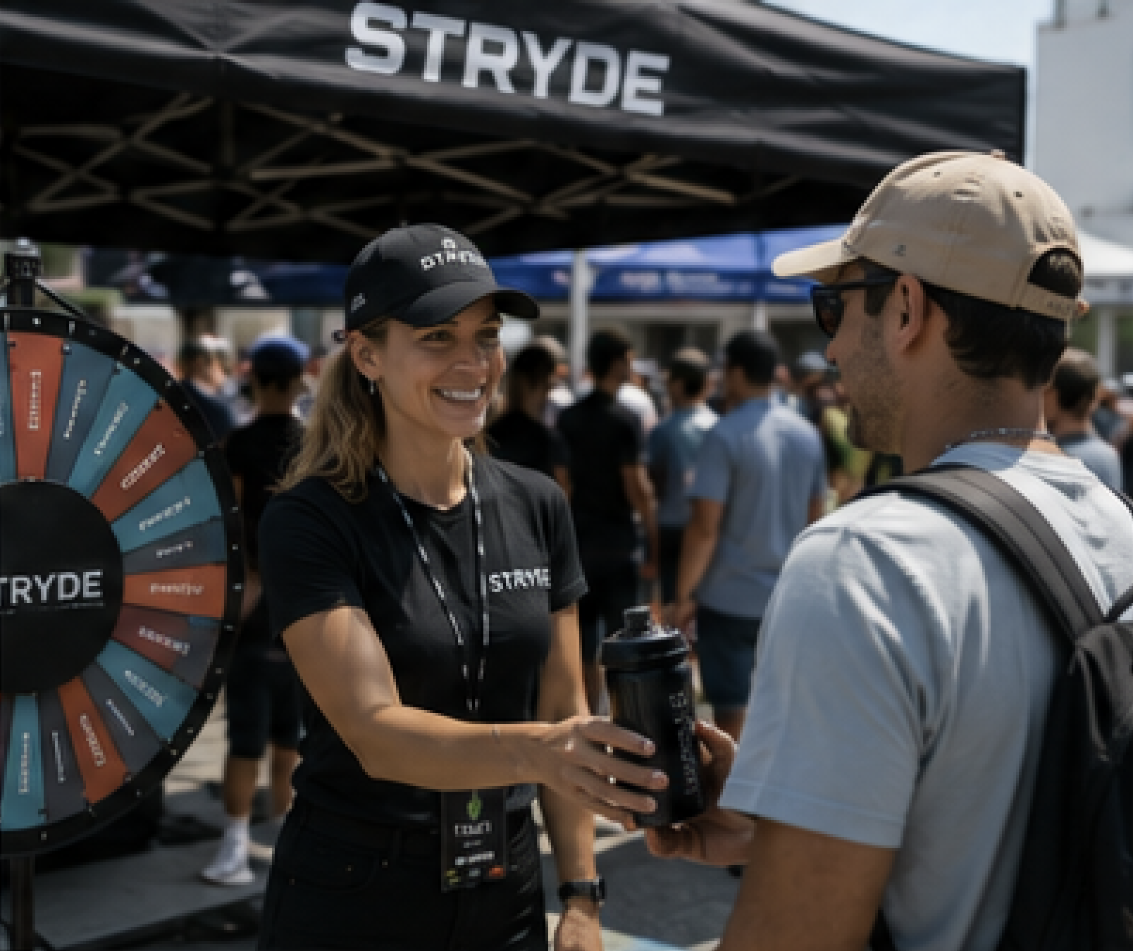 Brand ambassador in black uniform handing a drink to a guest beside a prize wheel at an outdoor activation